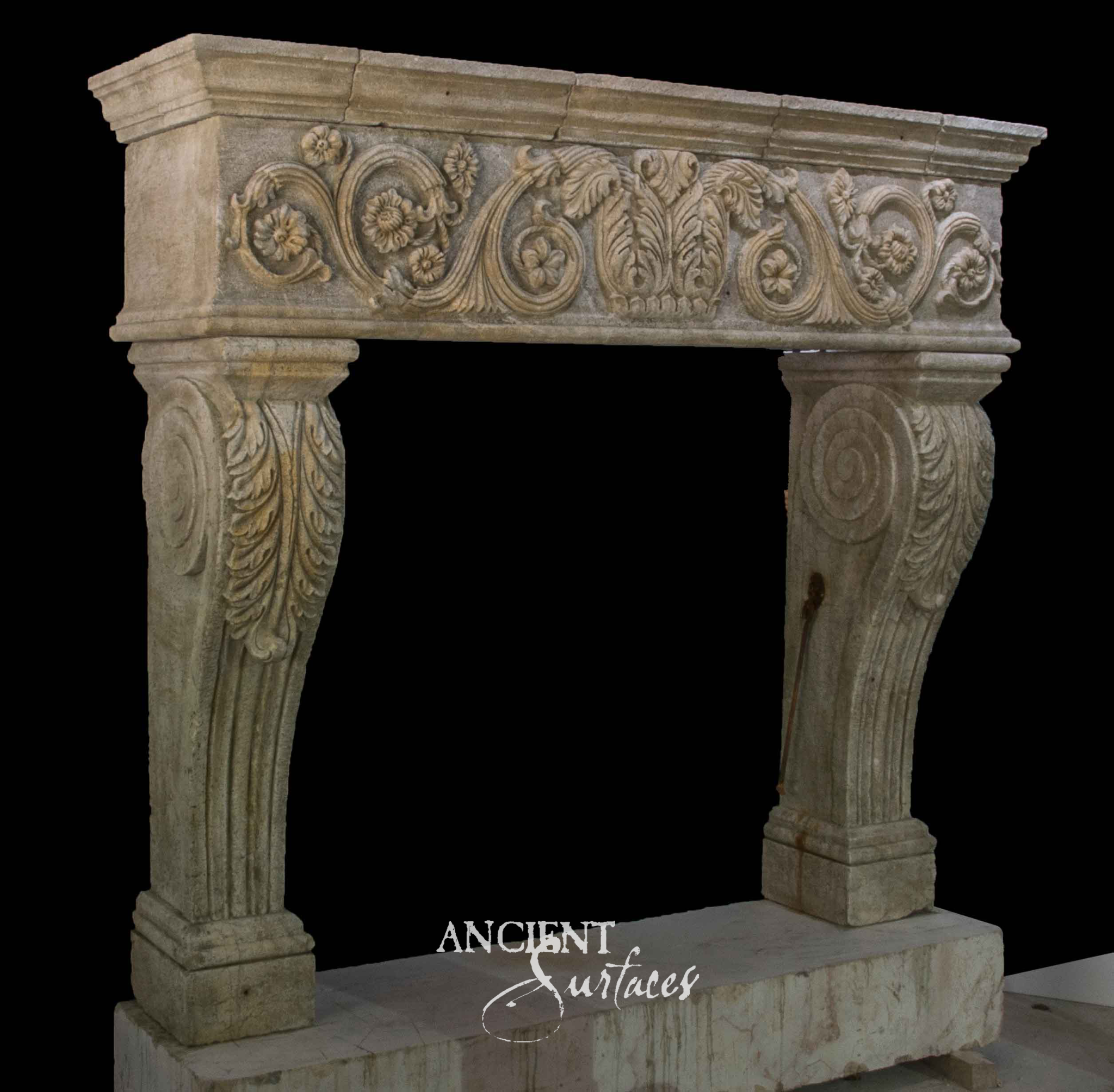 Reclaimed limestone fireplace mantel seen at an angle, with a scrolling blossom frieze and sweeping console jambs carved with acanthus leaves above fluted shafts
